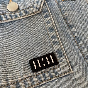 Puede incluir: Primer plano de una chaqueta vaquera azul claro con botones plateados y un pequeño parche negro. El parche tiene el texto blanco "11:11". La chaqueta tiene dos bolsillos delanteros y costuras visibles.