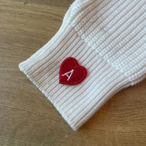 Puede incluir: Primer plano de una manga de punto color crema. Un parche rojo en forma de corazón con la letra blanca "A" está cosido en la manga. El tejido de punto tiene una textura acanalada.