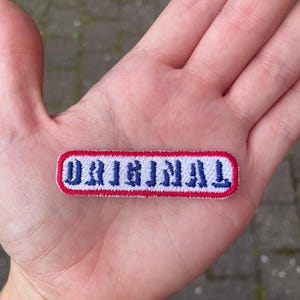 May include: Embroidered patch with the word "ORIGINAL" in blue, outlined in white, and framed by a red border. The patch is rectangular with rounded corners. The patch is held in a hand.