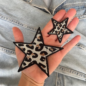 Op de afbeelding: Twee stervormige geborduurde patches met een luipaardprint in beige, bruin en zwart. Elke ster heeft een dikke, zwart gestikte rand die de vorm omlijnt. De patches worden in twee verschillende maten getoond, een grotere en een kleinere.