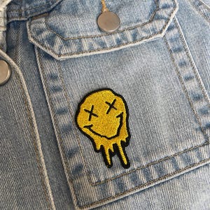 May include: A light blue denim jacket featuring a chest pocket with a silver metal button. An embroidered patch of a yellow melting smiley face with black stitched X-shaped eyes and a wide, dripping black mouth is attached to the pocket.