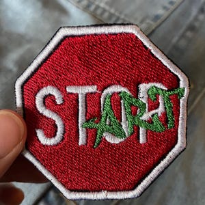May include: A red, white, and green embroidered patch in the shape of a stop sign. The word "STOP" is in white, and the word "ART" is in green, creating a playful design. The patch is likely for clothing or accessories.