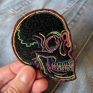 May include: Black skull patch with colourful embroidered details. The skull features a rainbow outline and internal accents in shades of red, yellow, green, and pink. The patch is held against a light blue denim background.