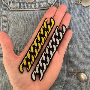 May include: Two long, narrow embroidered patches featuring a repeating chain link pattern. One patch is stitched in bright yellow thread on a black background, while the other is stitched in silver-grey thread on a black background, both with jagged edges.