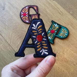 Może przedstawiać: Haftowane naszywki z literami w kolorze granatowym, bordowym i zielonym. Granatowa litera "A" jest w centrum uwagi, z haftem kwiatowym w kolorze białym, złotym i czerwonym. Bordowe "G" i zielone "P" są lekko rozmyte w tle, również z kwiatowymi wzorami.