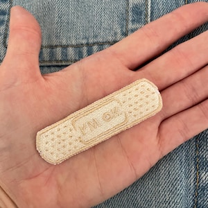 Puede incluir: Un parche de tela beige con forma de tirita, sostenido en una mano. El parche tiene una superficie texturizada con pequeños agujeros y el texto "YM OK" impreso. El fondo es una chaqueta vaquera.