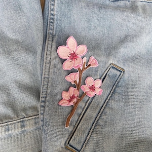 May include: A light blue denim jacket featuring an embroidered patch of a brown branch with several light pink cherry blossom flowers. The embroidery includes detailed dark pink centres and small buds, applied to the fabric near a pocket seam.