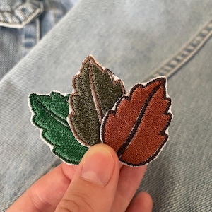 Op de afbeelding: Drie geborduurde bladpatches in groen, olijf en bruin met witte randen. De patches zijn gerangschikt in een cluster, met gedetailleerde stiksels en een naturalistisch ontwerp. Ideaal om een decoratief tintje toe te voegen aan kleding of accessoires.