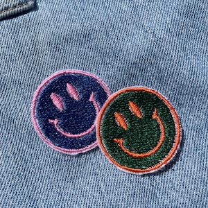 May include: Two embroidered smiley face patches on a blue denim background. One patch is navy blue with pink stitching and the other is green with orange stitching.