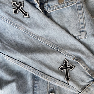 May include: A light blue denim jacket featuring two black and white embroidered cross patches on the sleeve. The jacket includes a classic collar, a button-down front with silver-toned metal buttons, and a chest pocket with a buttoned flap closure.