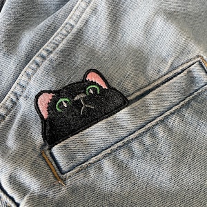 May include: A black cat patch with pink ears and green eyes peeks from a light blue denim pocket. The embroidered patch adds a playful touch to the denim. The denim has visible stitching and a classic, worn appearance.