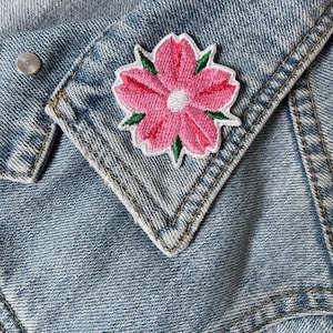 Può includere: Una toppa a forma di fiore rosa ricamato con foglie verdi e bordo bianco è applicata a una giacca di jeans blu chiaro. Il fiore ha un centro bianco e cuciture dettagliate.