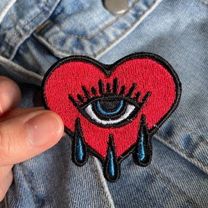 May include: Red heart-shaped embroidered patch with a blue eye and black eyelashes. The heart is dripping with blue teardrop shapes.