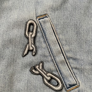 May include: Close-up of a light blue denim jacket with two chain patches and a rectangular patch. The chain patches are grey with black outlines. The rectangular patch is light blue with yellow stitching.