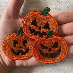 Könnte beinhalten: Drei gestickte Aufnäher mit orangefarbenen Kürbissen, grünen Stielen und schwarz gestickten Jack-o'-Lantern-Gesichtern. Jeder Kürbis zeigt einen anderen Gesichtsausdruck, darunter ein breites Grinsen, ein gezacktes Lächeln und ein bedrohlicher Blick.