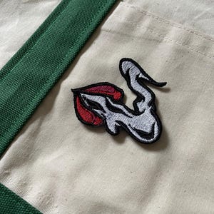 Peut inclure: Un sac fourre-tout beige avec une sangle verte et un écusson représentant un dessin stylisé de lèvres et de fumée. Les lèvres sont rouges et la fumée est blanche avec des contours noirs. L'écusson est cousu sur le sac.