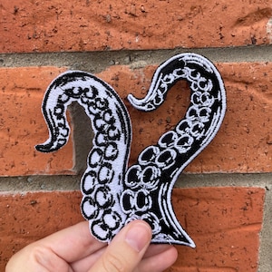 May include: An embroidered patch featuring two black and white octopus tentacles that curve upward. The design displays detailed circular suction cups along the inner length of each tentacle, with a thick black border outlining the entire shape.