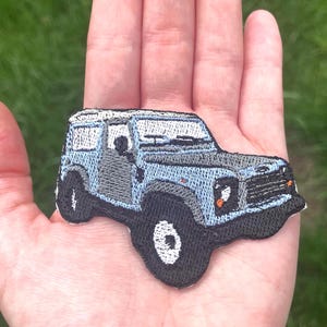 May include: An embroidered patch depicting a light blue and grey off-road vehicle. The patch has black tyres with white details and orange headlights. The vehicle design includes a detailed roof and windows, suitable for customising clothing or accessories.