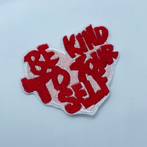 Könnte beinhalten: Gestickter Aufnäher in Herzform mit den Worten "BE KIND TO YOUR SELF" in roter, stilisierter Schrift. Der Aufnäher hat einen weißen Rand und einen hellrosa Hintergrund.