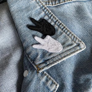 Könnte beinhalten: Zwei gestickte Patches, einer schwarz und einer weiß, auf eine Jeansjacke genäht. Beide Patches zeigen eine Hand, die das Friedenszeichen macht.