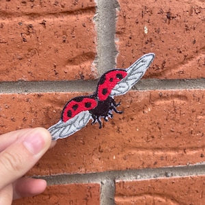 May include: An embroidered patch featuring a ladybug in flight with its wings spread wide. The ladybug has a black body, bright red wing covers with black spots, and light gray, textured wings extending outward to the sides.