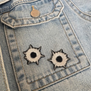 May include: A light blue denim jacket featuring a chest pocket with a silver-toned metal button. Two embroidered patches shaped like bullet holes, with black centers and jagged grey and white outlines, are stitched onto the front of the pocket.