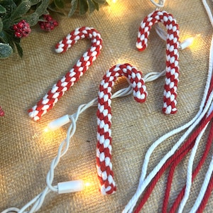 May include: Three red and white candy cane ornaments with a string of white lights. The candy canes are made of a woven material and are arranged on a textured surface with red and white string.