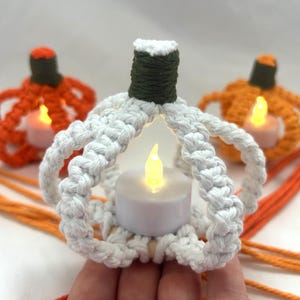 May include: Three macrame pumpkin candle holders in white, orange, and green. The white pumpkin is in focus, with a small LED candle inside. The orange pumpkins are in the background, also with LED candles. Orange yarn is in the foreground.