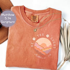 Peut inclure: T-shirt couleur terre cuite avec l'inscription "BE THE LIGHT" et un motif de coucher de soleil. Le texte "Matthew 5:14" est également présent. Un collier doré et un chapeau marron sont visibles.