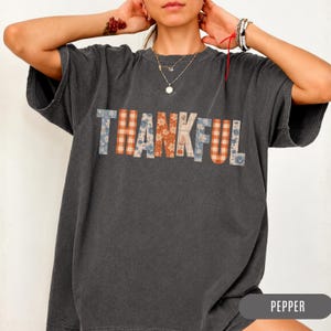 Peut inclure: Un t-shirt à manches courtes gris anthracite avec le mot THANKFUL imprimé sur la poitrine. Chaque lettre est remplie d'un motif différent, incluant des designs floraux bleus et orange ainsi que des carreaux vichy orange et blancs.