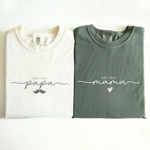 Peut inclure: Deux t-shirts pliés, un ivoire et un vert mousse. Le t-shirt ivoire porte le mot "papa" avec une moustache et "EST. 2025". Le t-shirt vert mousse porte le mot "mama" avec un cœur et "EST. 2025".