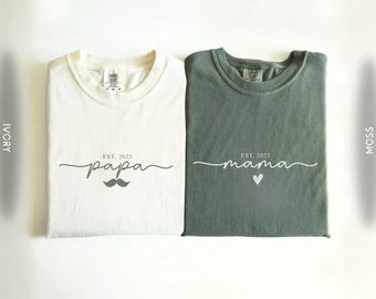 May include: Two folded t-shirts, one ivory and one moss green. The ivory shirt has the word "papa" with a mustache graphic and "EST. 2025". The moss green shirt has the word "mama" with a heart graphic and "EST. 2025".