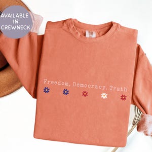 May include: Terracotta-colored long-sleeve shirt with the words "Freedom, Democracy, Truth" printed in white. Below the text are four small flower graphics in blue, red, white, and red. The shirt is available in a crewneck style.