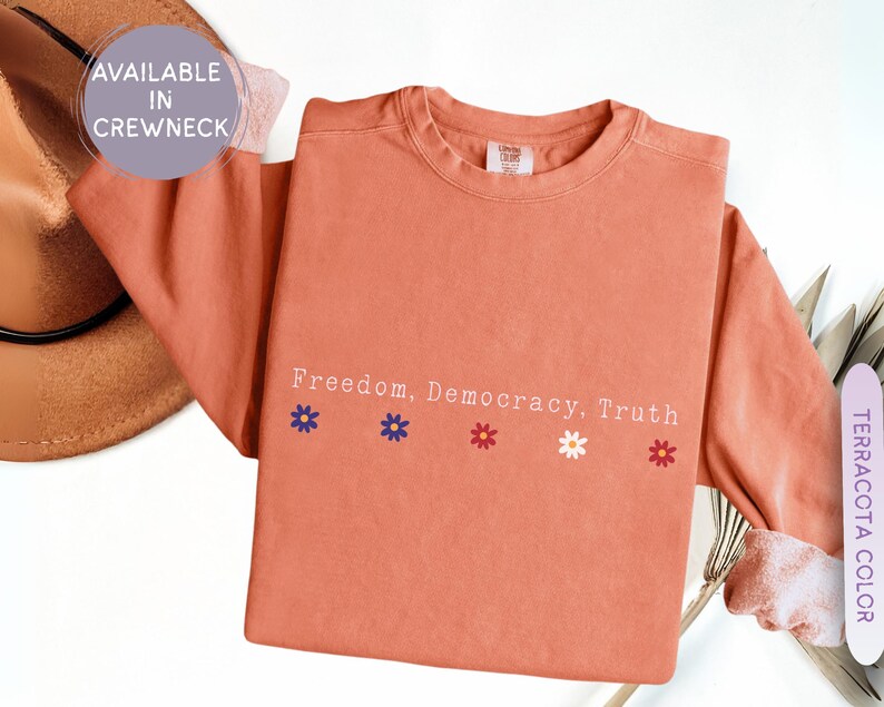 May include: Terracotta-colored long-sleeve shirt with the words "Freedom, Democracy, Truth" printed in white. Below the text are four small flower graphics in blue, red, white, and red. The shirt is available in a crewneck style.