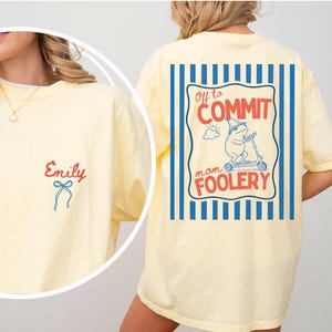 May include: A light yellow t-shirt with the name "Emily" in red script and a blue bow on the front. The back features a graphic of a frog on a scooter with the text "Off to Commit Mom Foolery" in a blue and red design.