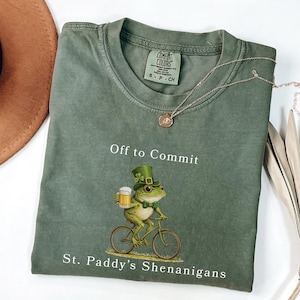 Peut inclure: T-shirt vert avec une grenouille portant un chapeau de leprechaun et tenant une chope de bière, à vélo. Le texte sur le t-shirt dit "Off to Commit St. Paddy's Shenanigans". Un collier doré et un chapeau marron sont également visibles.