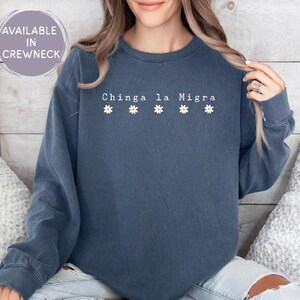 May include: A denim blue crewneck sweatshirt with the text "Chinga la Migra" and four small daisy graphics. The sweatshirt is a solid color and has long sleeves. The text is in white.