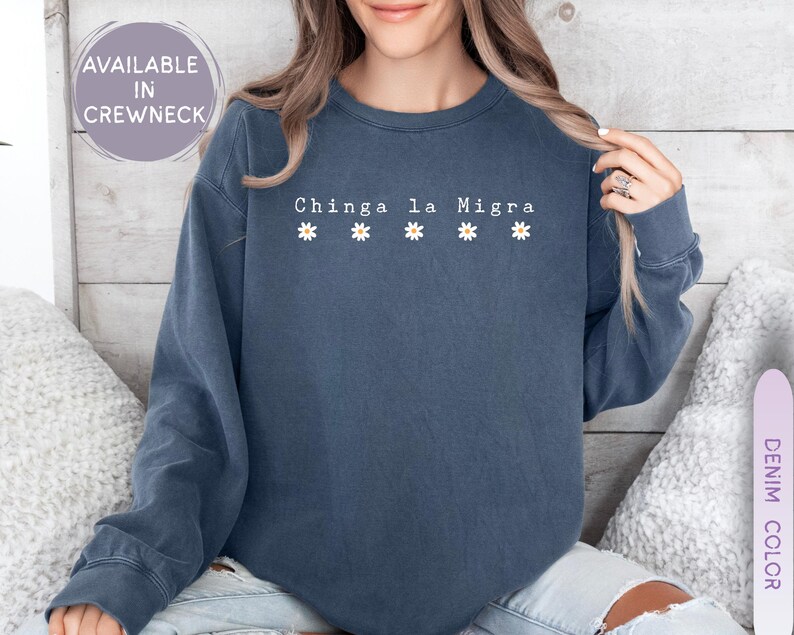 May include: A denim blue crewneck sweatshirt with the text "Chinga la Migra" and four small daisy graphics. The sweatshirt is a solid color and has long sleeves. The text is in white.