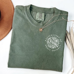 Peut inclure: Un t-shirt vert mousse avec l'inscription blanche "RADICALIZED BY BASIC DECENCY", accompagnée d'un motif d'oiseau et de fleurs. Un collier doré et un chapeau marron sont également visibles.