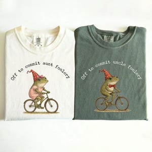 May include: Two t-shirts, ivory and moss green, with a whimsical frog design. The ivory shirt features a frog in a dress riding a bicycle, with the text "Off to commit aunt foolery." The moss green shirt has a frog in a bow tie riding a bicycle, with the text "Off to commit uncle foolery."