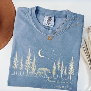 May include: A blue short-sleeve t-shirt featuring a cream-colored graphic of a forest, a crescent moon, stars, and two bears. The text reads "Mama bear" followed by "Emily &hearts; Oliver". The shirt has a Comfort Colors brand label at the neck.
