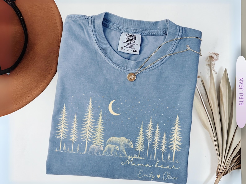 May include: A blue short-sleeve t-shirt featuring a cream-colored graphic of a forest, a crescent moon, stars, and two bears. The text reads "Mama bear" followed by "Emily &hearts; Oliver". The shirt has a Comfort Colors brand label at the neck.