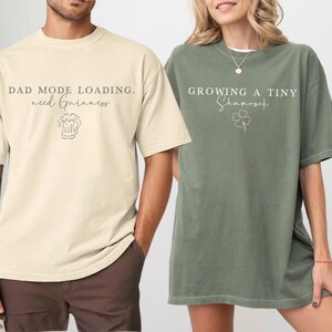 May include: Two oversized t-shirts. One is ivory with the text "DAD MODE LOADING. need Guinness" and a beer graphic. The other is moss green with the text "GROWING A TINY Shamrock" and a shamrock graphic. Both shirts have short sleeves.
