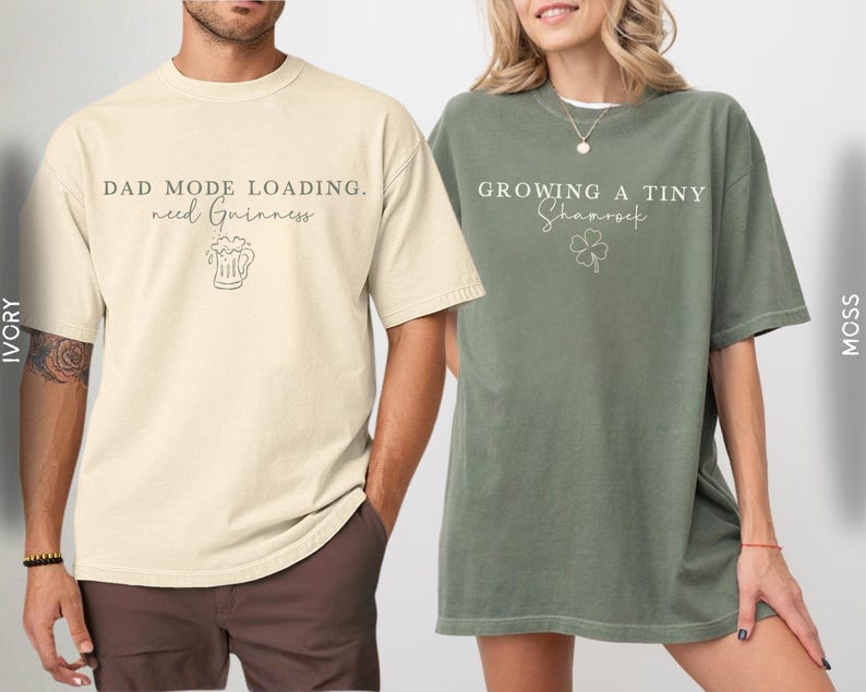 May include: Two oversized t-shirts. One is ivory with the text "DAD MODE LOADING. need Guinness" and a beer graphic. The other is moss green with the text "GROWING A TINY Shamrock" and a shamrock graphic. Both shirts have short sleeves.