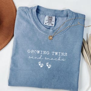 Peut inclure: Un t-shirt bleu jean avec le texte "GROWING TWINS send snacks" en écriture blanche. Le t-shirt comporte deux paires d'empreintes de pas de bébé en dessous. Un collier doré avec un pendentif repose sur le t-shirt.