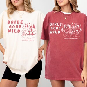 Puede incluir: Dos camisetas, una marfil y otra carmes&iacute;, con los textos "BRIDE GONE WILD" y "GIRLS GONE WILD". Cada camiseta presenta un gr&aacute;fico con tem&aacute;tica de camping y el texto "Emily's Bachelorette 2025 Lake Tahoe, CA."