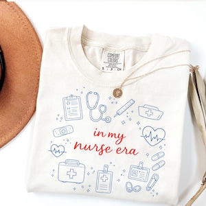 Peut inclure: T-shirt crème avec l'inscription "in my nurse era" en rouge, entourée d'illustrations bleues sur le thème médical. Un collier doré et un chapeau marron sont également visibles.