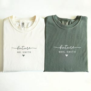 May include: Two folded t-shirts, one ivory and one moss green, with the word "future" in script. The ivory shirt says "Mr. Smith" and the green shirt says "Mrs. Smith", both with a heart symbol below. The shirts are likely for a couple.