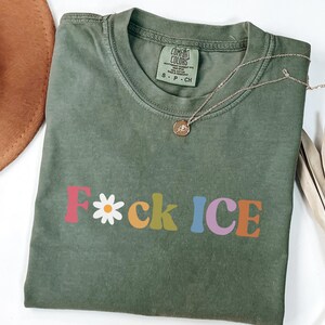 May include: Olive green t-shirt with the text "F*ck ICE" in colorful, retro-style lettering. A daisy replaces the "o" in "F*ck". A gold necklace with a pendant rests on the shirt. A brown hat is visible in the background.
