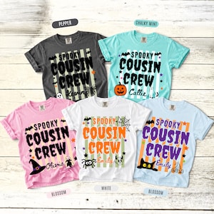 Peut inclure: Cinq t-shirts à manches courtes en gris anthracite, menthe, rose, blanc et bleu clair, chacun avec un fond à rayures verticales et le texte "SPOOKY COUSIN CREW" accompagné de motifs d'Halloween comme des fantômes, citrouilles et araignées.